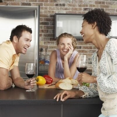 Three people sit at a kitchen counter with wine glasses, bread, and vegetables—smiling and chatting as if checking off an impromptu party supply list together.