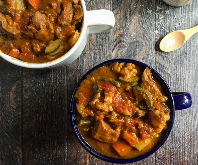 beef stew in a big blue mug and a big white mug on a brown table next to a wooden spoon
