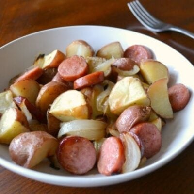 A white bowl filled with sliced roasted potatoes, sausage, and onions from a delicious sausage onion and potato sheet pan dinner sits on a wooden table with a fork in the background.