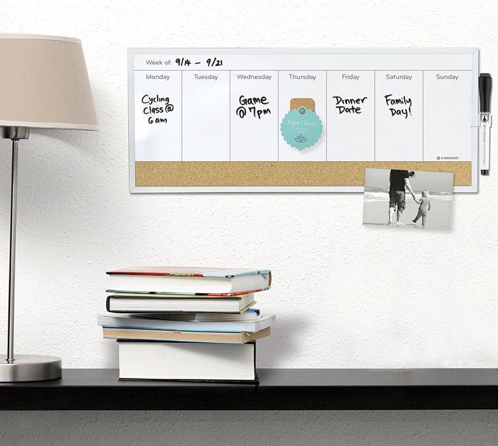a table with a lamp and a stack of books on it with a weekly whiteboard calendar on the wall behind it