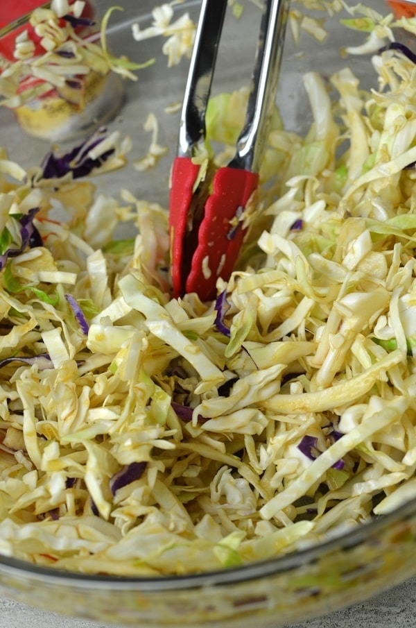 tongs being used to toss coleslaw mixture in a glass bowl