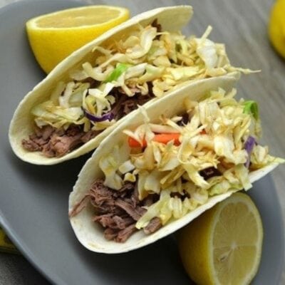 Two Asian tacos filled with shredded meat and cabbage slaw are served on a gray plate alongside lemon halves.