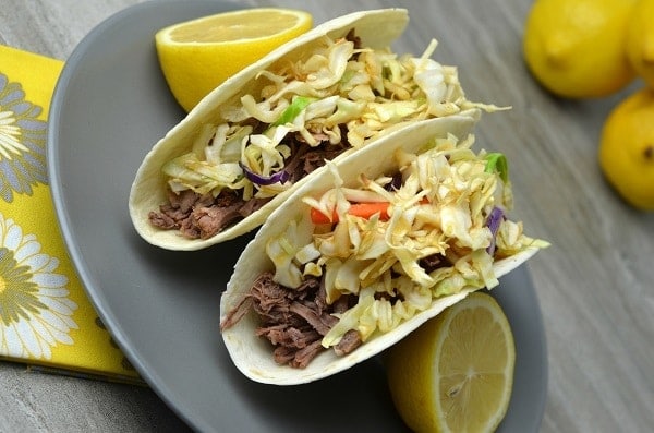 Asian Tacos next to lemon halves on a gray plate on a yellow flower cloth on a gray table