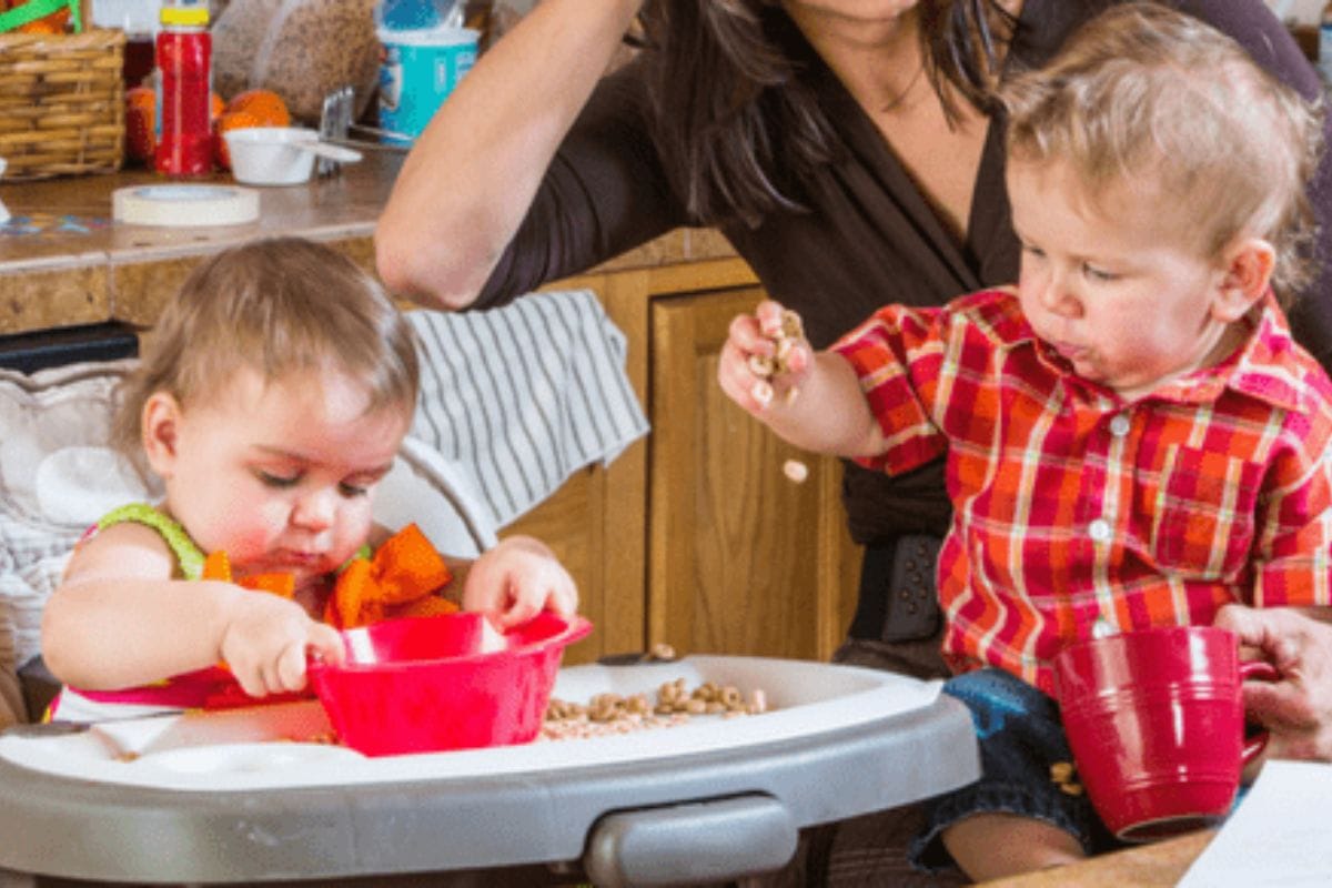 Two toddlers sit in high chairs at a kitchen counter, playing with food in red bowls and cups. An adult sits behind them, mostly out of frame-perfect moment for trying out cleaning hacks for moms after mealtime fun.