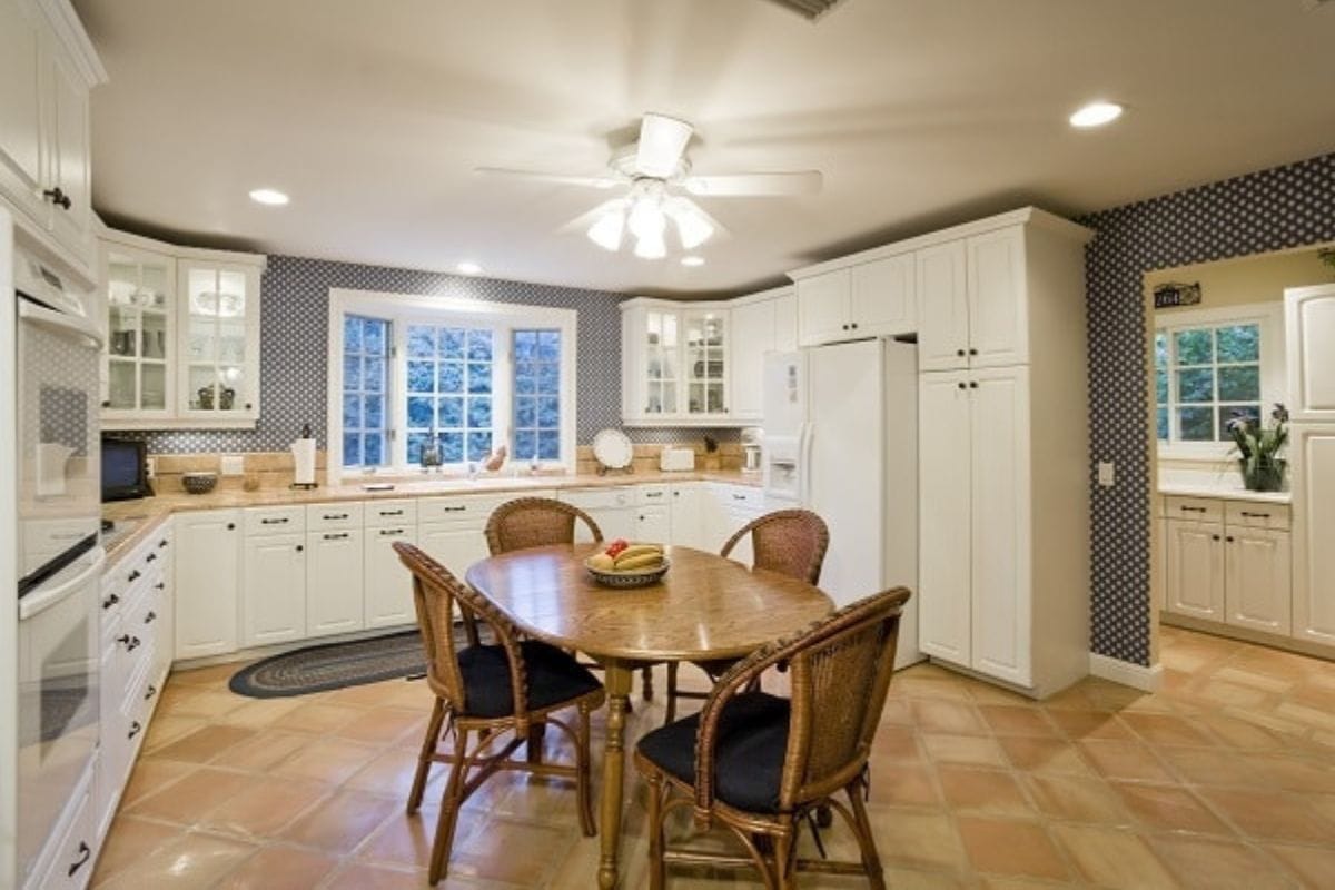 A spacious kitchen with white cabinets, a round wooden table with four wicker chairs, tiled floor, and a ceiling fan with lights-perfect for trying out cleaning hacks for moms.