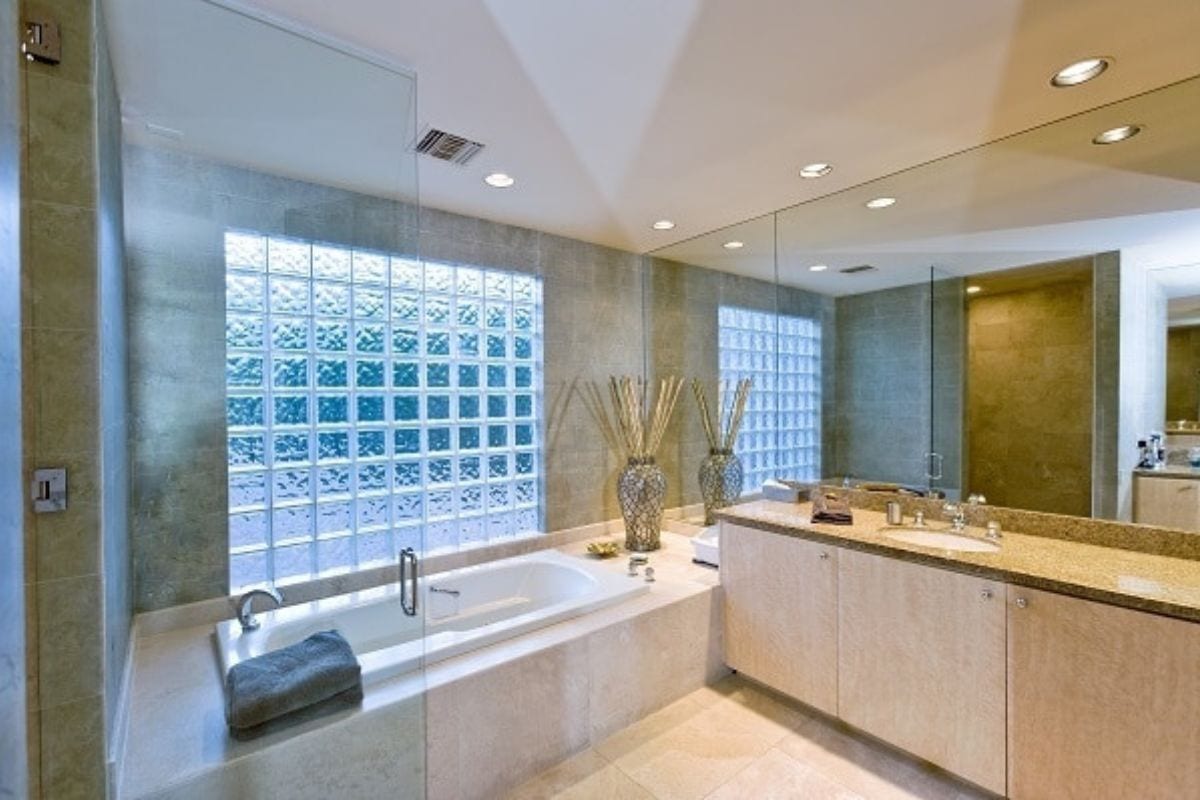 Modern bathroom with a large bathtub, glass block window, beige tiled walls, double sink vanity, and decorative vases on the counter-perfect for trying out new cleaning hacks for moms.