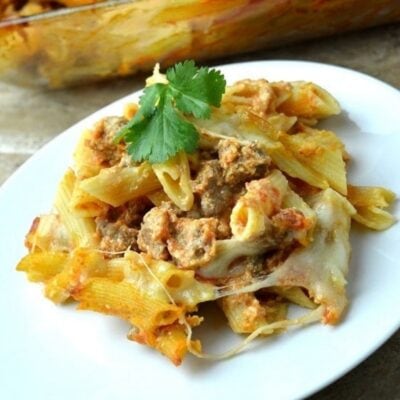 Baked penne pasta with ground meat, tomato sauce, and melted cheese, reminiscent of a lasagna casserole, garnished with a sprig of cilantro on a white plate.