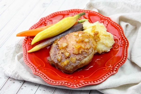 Maple Glazed Baked Pork Chops next to vegetables and potatoes on a red plate on a white cloth on a white table