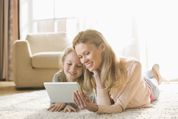 a mother and daughter laying on the carpet looking at an ipad