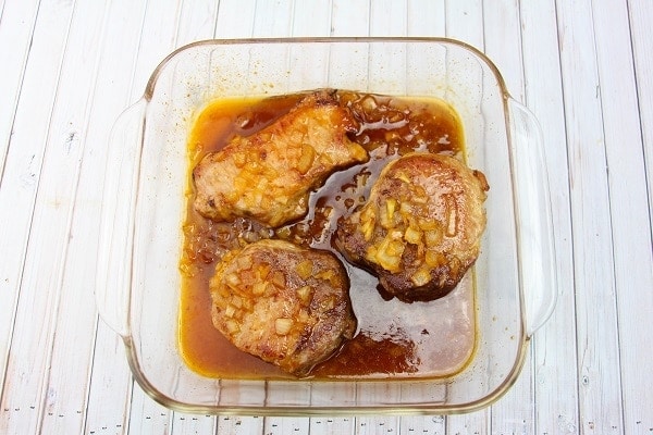 pork chops covered with sauce in a glass baking dish on a white wood table