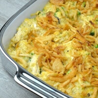 A close-up of a chicken broccoli quinoa casserole topped with crispy fried onions, showing a mixture of baked vegetables and chicken in a white baking dish.