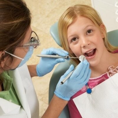 A dentist examines a young girl's teeth using dental instruments while she sits in a dental chair, wearing a bib, ensuring children's dental health is maintained.