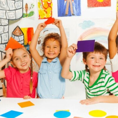 Four children sit at a table holding up colored paper shapes and smiling, with artwork and drawings displayed on the wall behind them—creating a cheerful scene perfect for indoor party games.