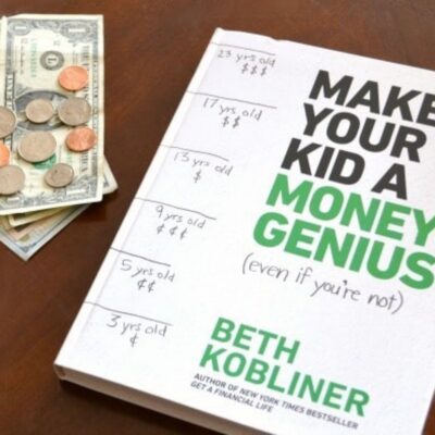 A book titled "Make Your Kid a Money Genius" by Beth Kobliner sits next to a stack of cash and coins on a wooden surface, highlighting smart ways to make your kid a money genius. Handwritten notes with ages appear on the book cover.