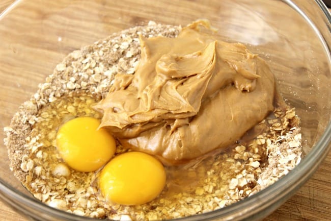 dry ingredients for No Flour Peanut Butter Protein Cookies and eggs, vanilla and peanut butter in a glass mixing bowl on a brown table