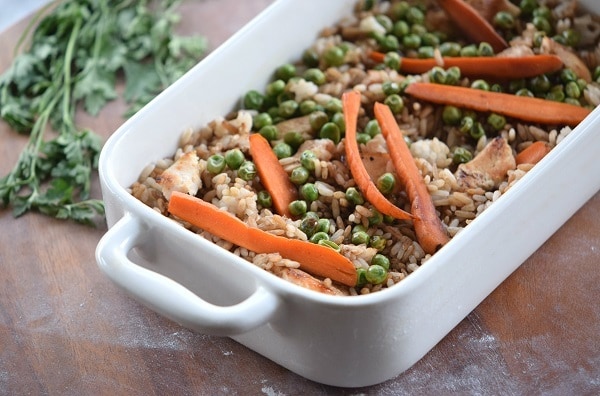 teriyaki chicken with rice and peas topped with carrots in a white baking dish on a brown table next to parsley