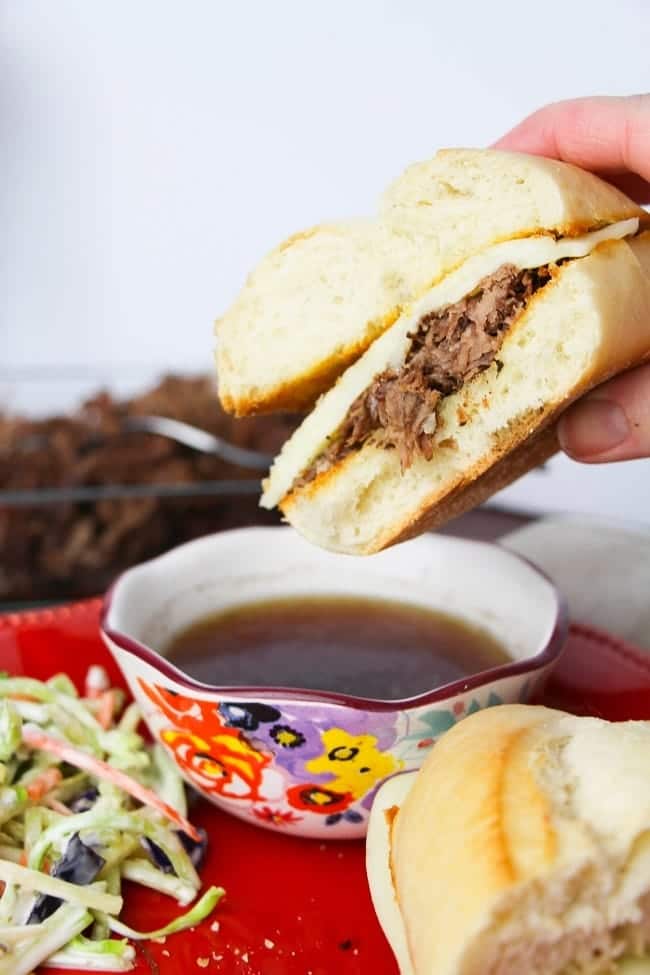 a hand holding a french dip sandwich on a red plate with sauce in a bowl