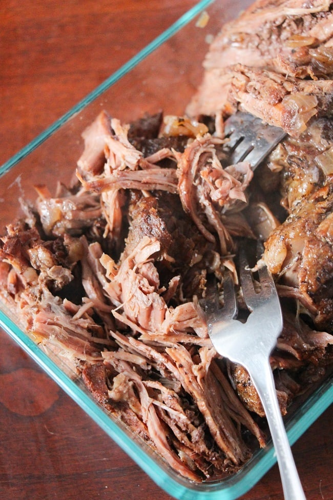 the beef roast in a glass dish being shredded with two forks