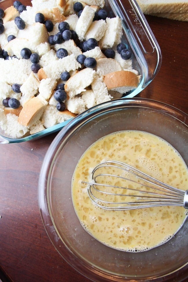 egg mixture in a glass bowl with a whisk in it next to cubed bread and blueberries in a glass baking dish, all on a brown table