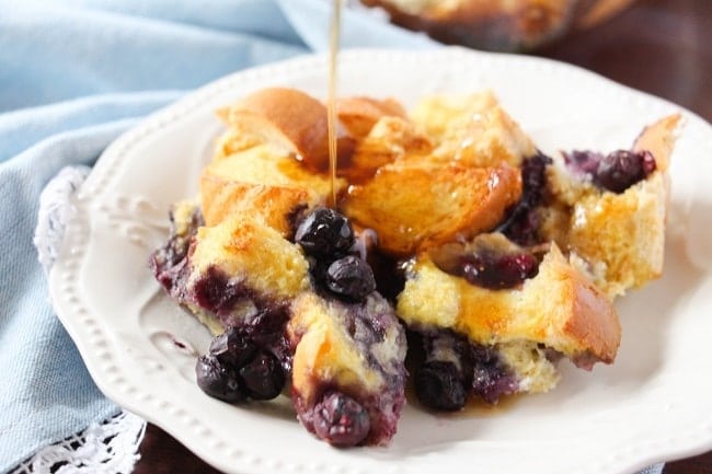 make ahead blueberry french toast breakkfast casserole on a white plate with syrup being poured on it, near a blue linen on a brown table