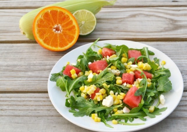 sweet corn and watermelon salad with citrus dressing on a white plate on a wood table with an orange and lime slice, and corn on the cob in the background
