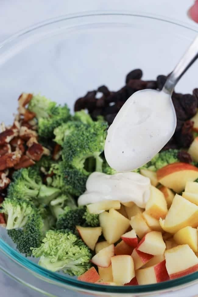 a glass bowl of chopped broccoli, apples, nuts and raisins on a white table being topped with dressing from a spoon
