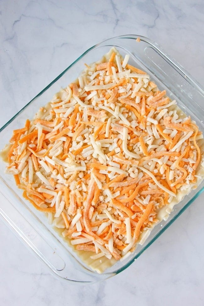 lasagna noodles, filling and shredded cheese in a glass dish on a white marbled counter