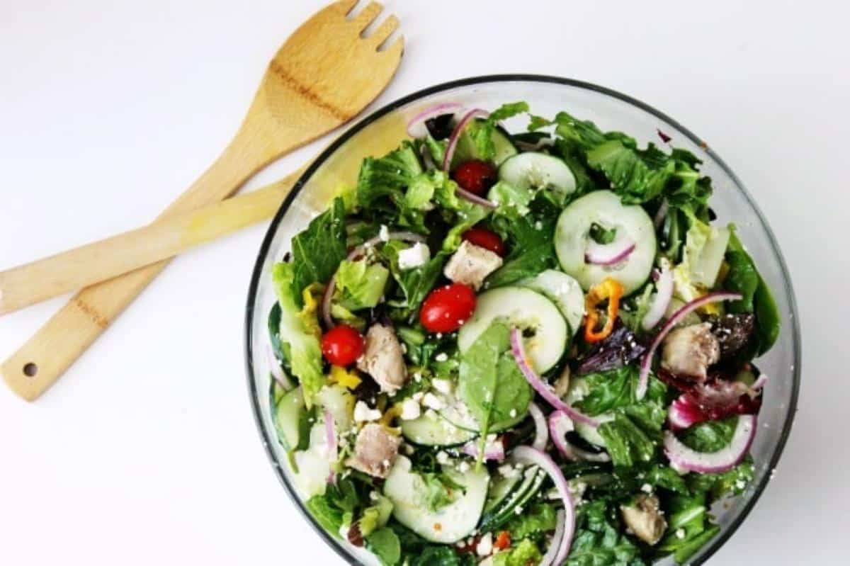 Overhead view of Greek salad in a glass bowl next to a wooden spoon and fork on a white background.