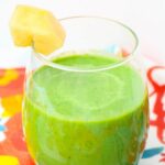 A glass of cantaloupe green smoothie with a pineapple chunk garnish on the rim, set on a colorful surface.