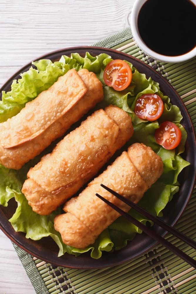 fried chicken spring rolls on top of lettuce next to sliced tomatoes on a black plate with black chopsticks on it, on a bamboo mat next to a white bowl of sauce 