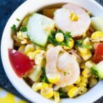A bowl of shrimp salad with grilled corn, cucumber, tomato, and parsley, all mixed together in a white dish.