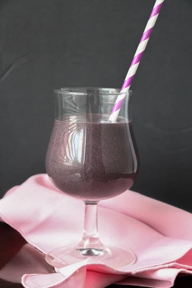 blueberry spinach smoothie in a glass with a pink and white straw in it, on a pink cloth with a black background