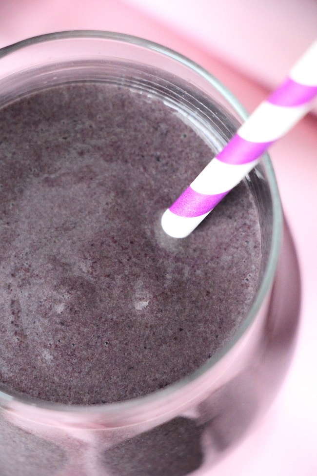 an overhead closeup view of a blueberry smoothie in a glass with a pink and white striped straw in it on a pink cloth 
