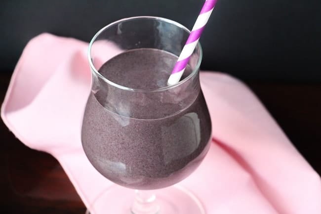 a blueberry smoothie in a glass with a pink and white striped straw in it on a pink cloth with a dark background 