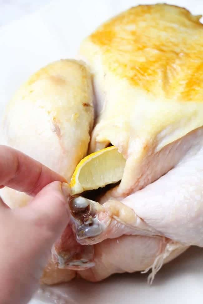 a hand putting a lemon slice into the cavity of a whole chicken