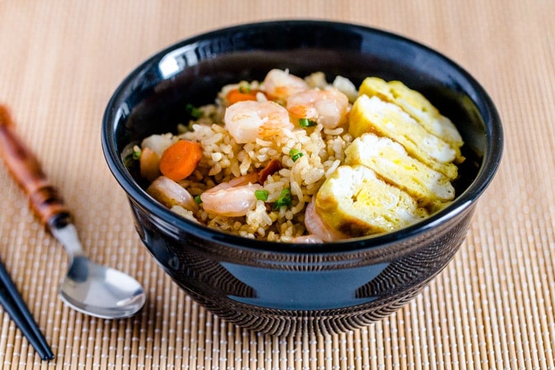 Japanese fried rice with shrimp in a black bowl next to a spoon on a bamboo mat