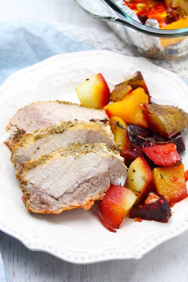 roasted garlic and herb pork loin, potatoes, beets and squash on a white plate with a glass baking dish with more pork and vegetables in it in the background, all on a white table