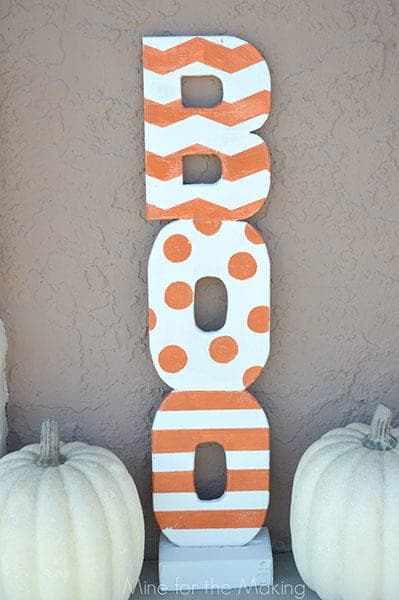 wooden letters painted orange and white to spell out the word Boo, next to two white pumpkins against the outside wall of a house