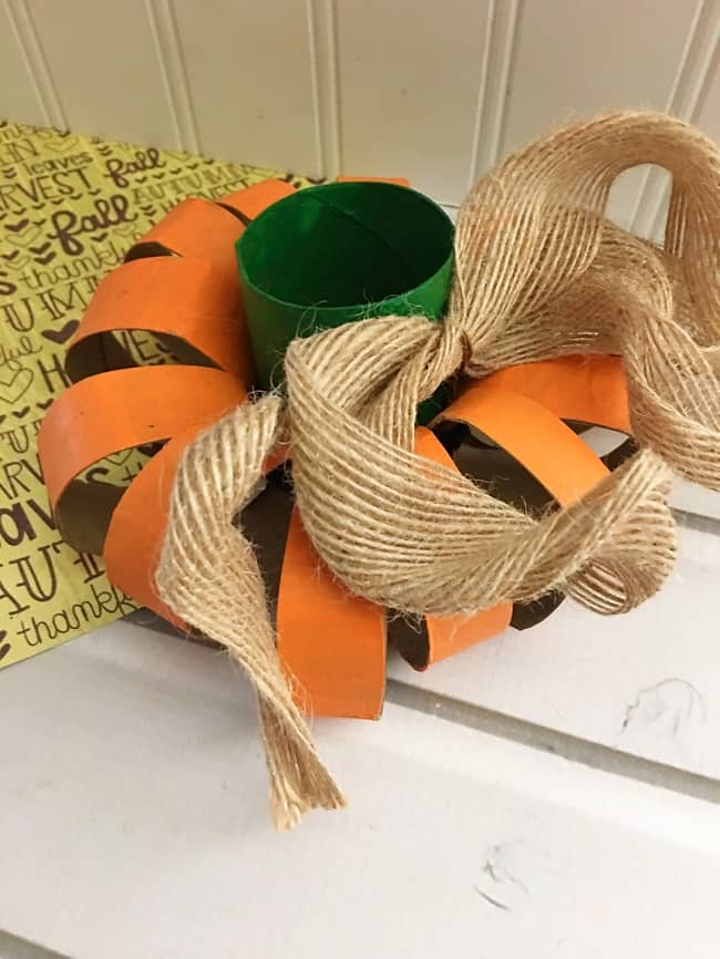 orange cardboard rings with a green toilet paper roll in the center next to a burlap ribbon on a white wood background