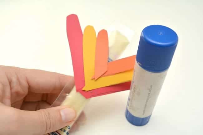 a hand holding a cheese stick with orange, yellow and red paper glued to it to look like turkey feathers, next to a glue stick on a white background
