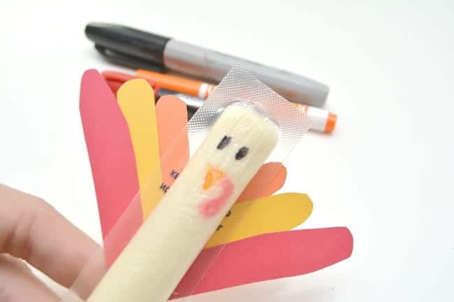 a hand holding a cheese stick with orange, yellow and red paper glued to it to look like turkey feathers with a face drawn on it with markers that are in the background, all on a white background