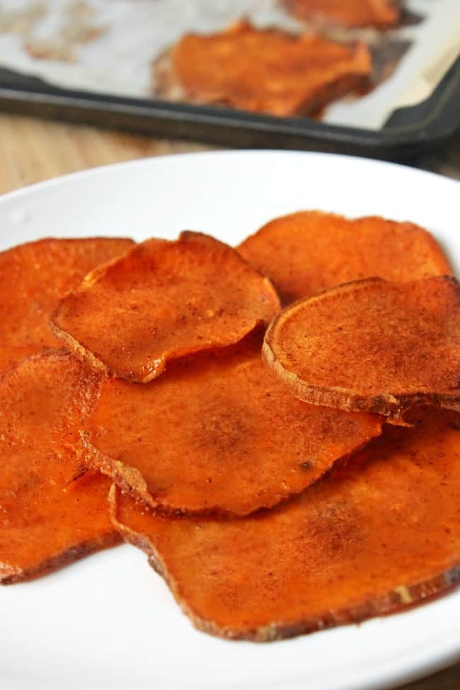 sliced sweet potatoes coated with cinnamon sugar on a plate with more in the background on parchment paper on a baking sheet