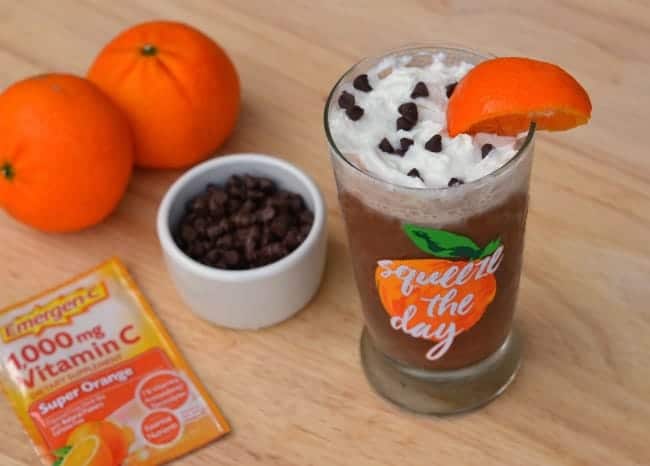 orange frappuccino topped with whipped cream, mini chocolate chips and an orange wedge in a glass with words on it reading squeeze the day next to two oranges, a white bowl of mini chocolate chips, a package of Emergen-C vitamin c, all on a wood table