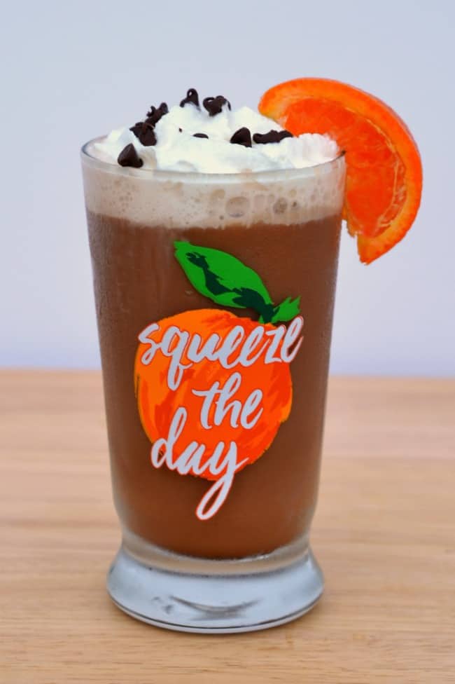 orange frappuccino topped with whipped cream, mini chocolate chips and an orange wedge in a glass with words on it reading squeeze the day on a wood table