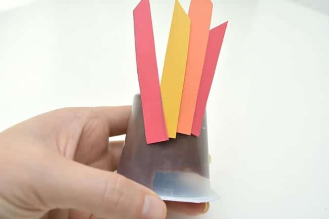 a hand holding an upside down pudding cup with red, yellow and orange paper feathers on it