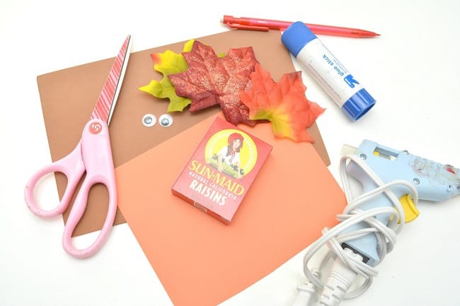 orange cardstock, fabric leaves in fall colors, scissors, a pencil, glue gun, googly eyes, and a box of raisins all on a white background