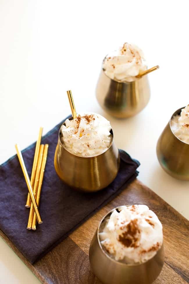 pumpkin spice cocktail topped with whipped cream in four metal cups on a dark linen, on a brown piece of wood, and on a white table
