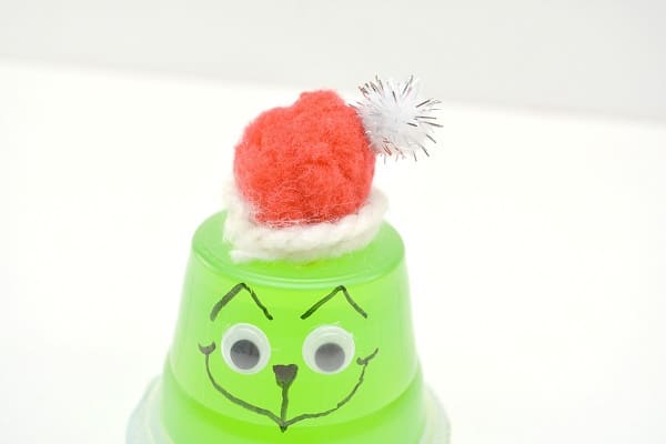 an upside down green jello cup with google eyes on it, a face drawn on with a black sharpie, topped with a red and white pom pom wrapped in white yarn to look like the grinch on a white background