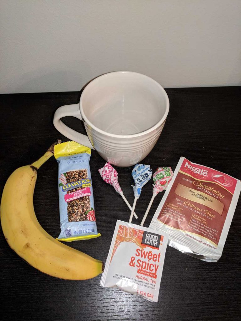 a white mug, banana, protein bar, lollipops, hot cocoa mix, tea on a brown table