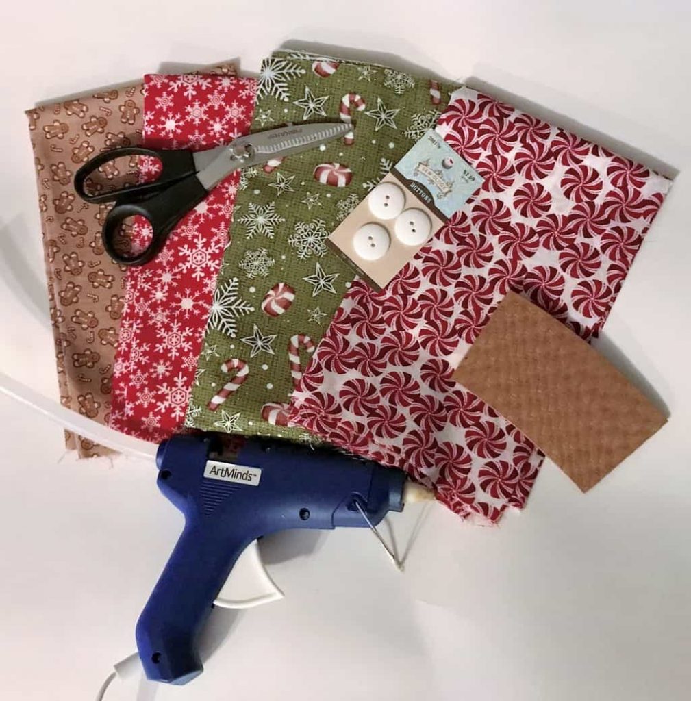 four different types of Christmas fabric, scissors, white buttons, a brown paper coffee holder, and a glue gun on a white background